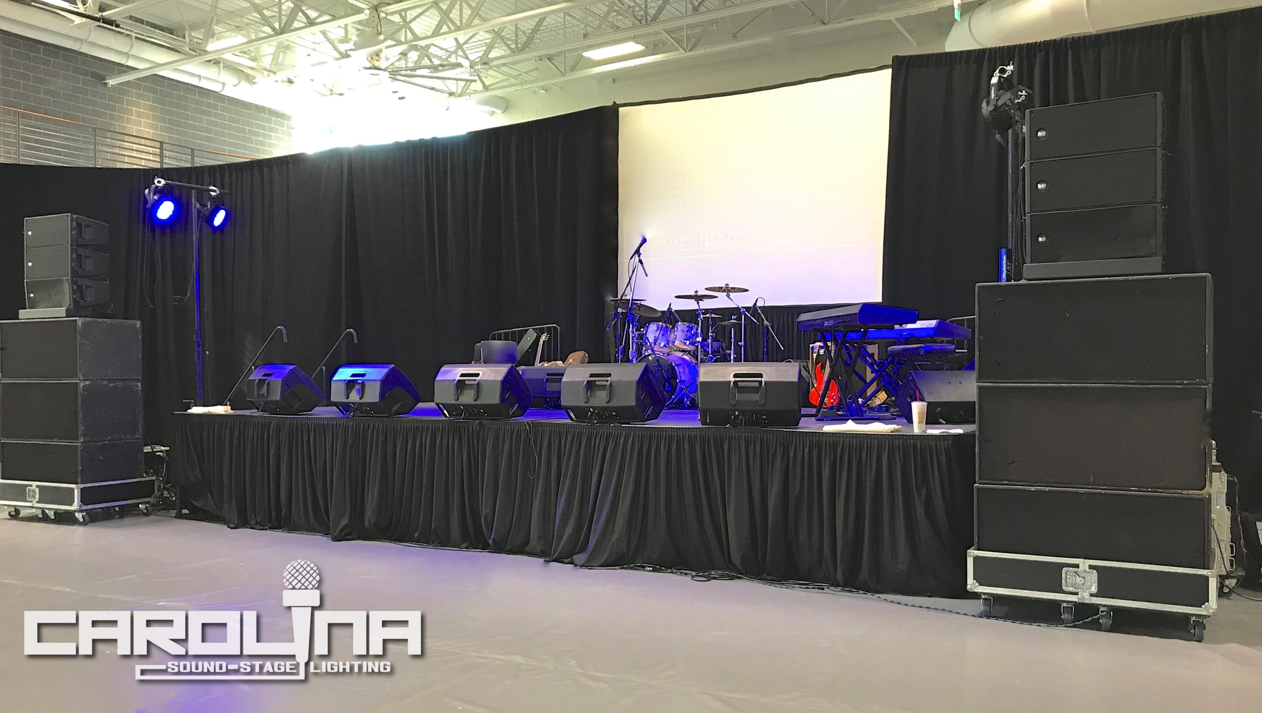 24'x16' stage equipped with RCF/JBL audio, black velour pipe & drape backdrop, rear video projection screen, band backline, and basic LED stage wash lighting
